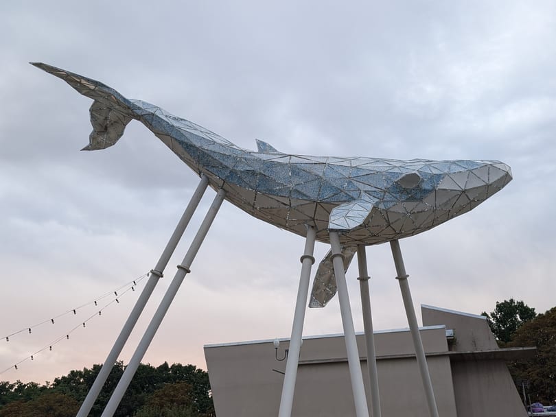 The Kyiv Whale, a faceted sculpture of a whale raised on white metal poles, seen from below against a pale evening sky