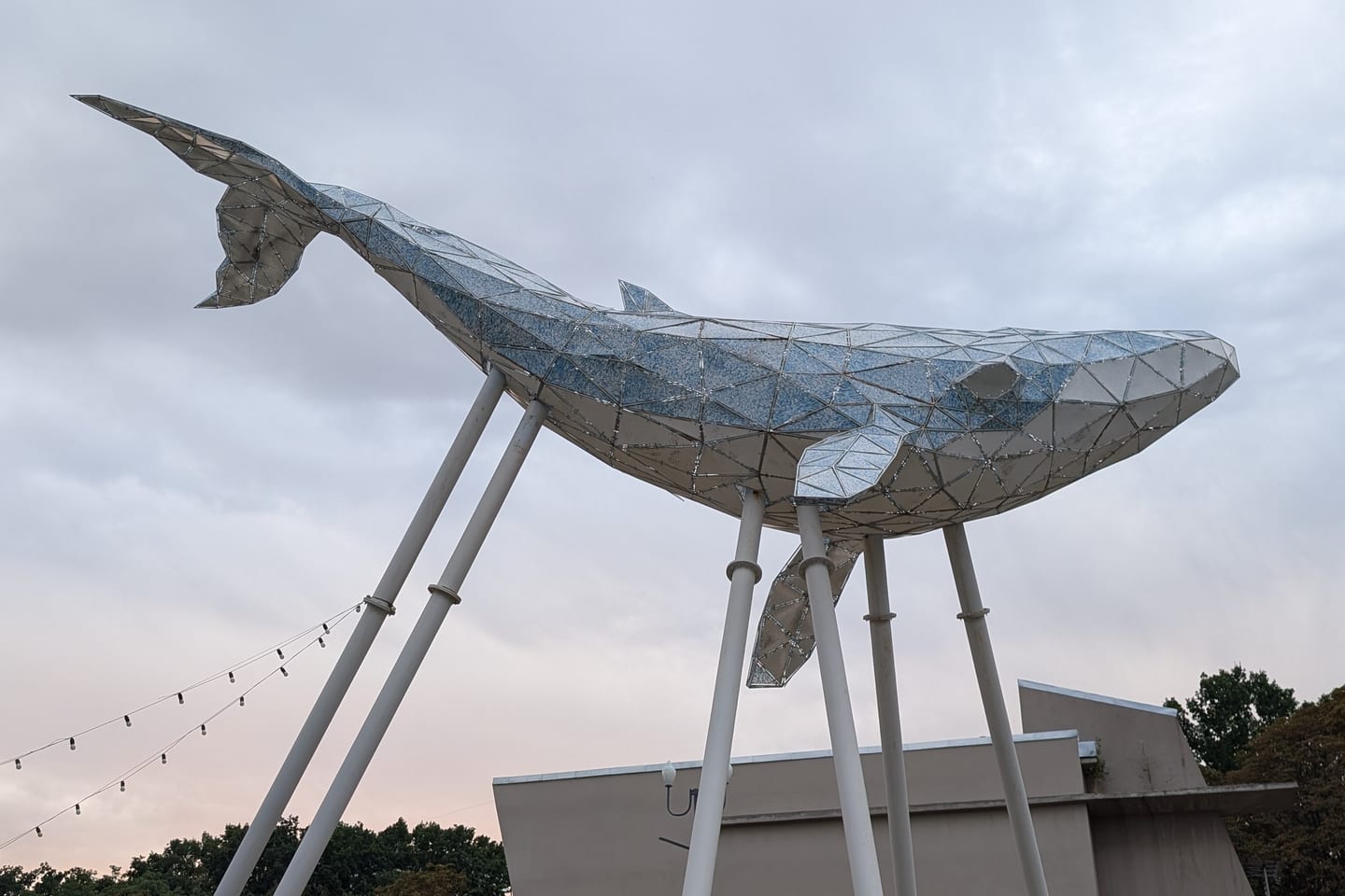 The Kyiv Whale, a faceted sculpture of a whale raised on white metal poles, seen from below against a pale evening sky