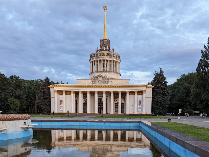 The main pavilion at VDNH, a white colonnaded building with a gold spire topped by a red star, reflected in the fountain pool in front of it