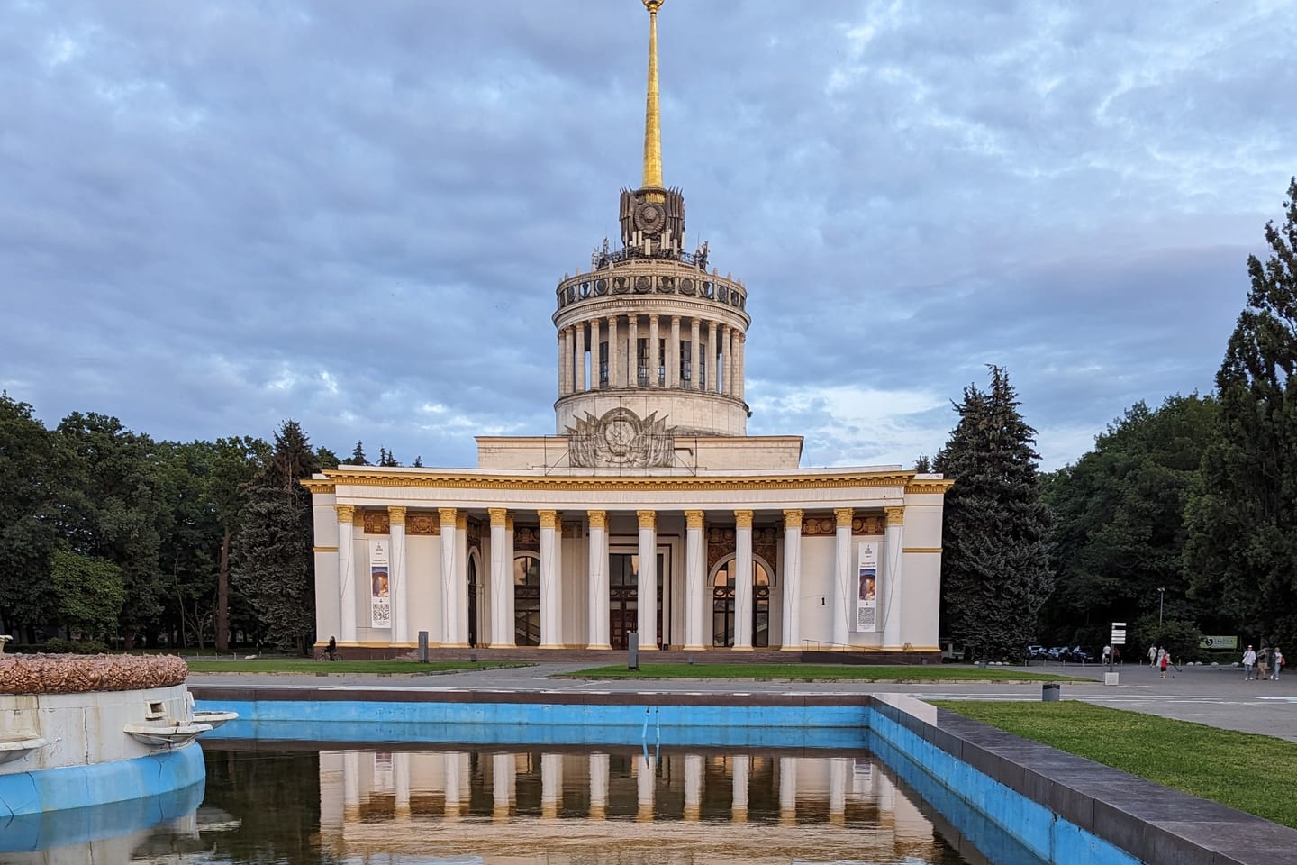 The main pavilion at VDNH, a white colonnaded building with a gold spire topped by a red star, reflected in the fountain pool in front of it